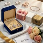 Das generierte Bild zeigt eine luxuriöse Geschenk-Szene auf weißem Marmortisch: Im Zentrum eine geöffnete elegante Schmuck Geschenkbox aus Samt in Dunkelblau mit goldenen Details, in der eine funkelnde Halskette mit Herzanhänger und passende Ohrringe auf weißem Satin liegen. Daneben mehrere Schmuck Geschenksets in verschiedenen Größen – eine Roségold-Box mit Schleife, eine moderne minimalistische weiße Box und eine vintage-inspirierte Schatulle. Im Vordergrund liegen dekorativ arrangiert: Geschenkbänder in Gold und Rosa, eine kleine Karte mit "For You" Aufschrift, frische Rosen in Pastelltönen und ein personalisierter Anhänger mit Gravur. Im Hintergrund sanft unscharf weitere Schmuckstücke – Perlenohrringe, Stapelringe und ein Armband. Die warme, natürliche Beleuchtung mit goldenem Schimmer erzeugt eine festliche, emotionale Atmosphäre – perfekt für einen Artikel über Schmuckgeschenke.