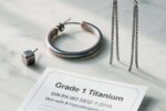 Das Bild zeigt eine elegante Produktpräsentation verschiedener Titanohrringe auf weißem Marmor: Minimalistische Ohrstecker, moderne Creolen und hängende Designs in natürlichem Silbergrau sowie anodisierten Roségold- und Blautönen. Im Vordergrund liegt ein medizinisches Zertifikat mit "Grade 1 Titanium" Aufschrift. Die Komposition vermittelt Reinheit, Qualität und Hautverträglichkeit .
