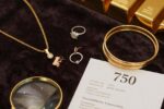 Das Bild zeigt eine luxuriöse Produktpräsentation verschiedener Goldschmuckstücke auf dunklem Samt: Eine elegante 18-Karat-Gelbgoldkette, Roségold-Ohrringe, ein Weißgold-Ring mit Diamant und gestapelte Goldarmbänder in verschiedenen Karatierungen. Im Vordergrund liegt eine Juwelierlupe neben einem Echtheitszertifikat mit sichtbarer "750"-Punzierung. Goldbarren im Hintergrund symbolisieren die Wertanlage. Die warme, professionelle Beleuchtung betont den Glanz und die Qualität des Goldes – perfekt für einen Kaufratgeber.
