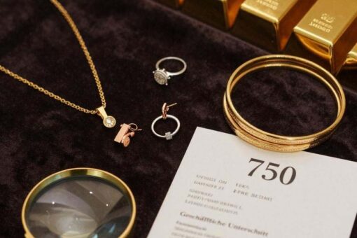 Das Bild zeigt eine luxuriöse Produktpräsentation verschiedener Goldschmuckstücke auf dunklem Samt: Eine elegante 18-Karat-Gelbgoldkette, Roségold-Ohrringe, ein Weißgold-Ring mit Diamant und gestapelte Goldarmbänder in verschiedenen Karatierungen. Im Vordergrund liegt eine Juwelierlupe neben einem Echtheitszertifikat mit sichtbarer "750"-Punzierung. Goldbarren im Hintergrund symbolisieren die Wertanlage. Die warme, professionelle Beleuchtung betont den Glanz und die Qualität des Goldes – perfekt für einen Kaufratgeber.